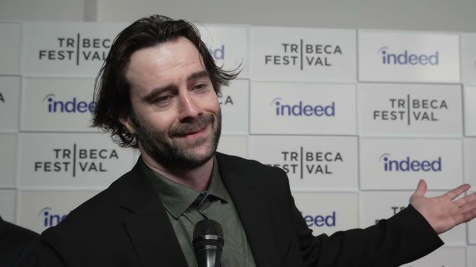 Ritratto ravvicinato di Cillian O'Sullivan al Tribeca Festival, che indossa un completo scuro e una camicia verde-grigio, mentre parla in un microfono.