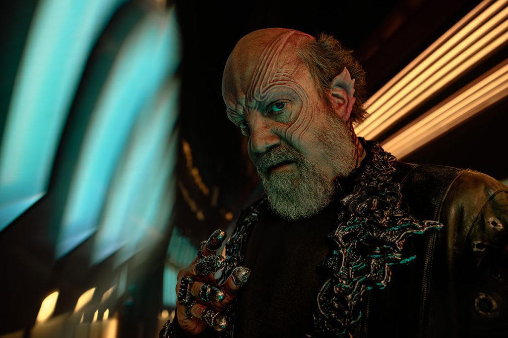 Paul Giamatti come Nus Braka, un alieno calvo con rughe profonde, barba grigia, orecchie a punta, indossa un giubbotto di pelle scuro e anelli elaborati.