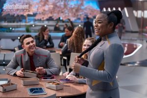 Kerrice Brooks sorride tenendo le sue trecce in una mensa futuristica con altri studenti, tra cui George Hawkins e Bella Shepard.