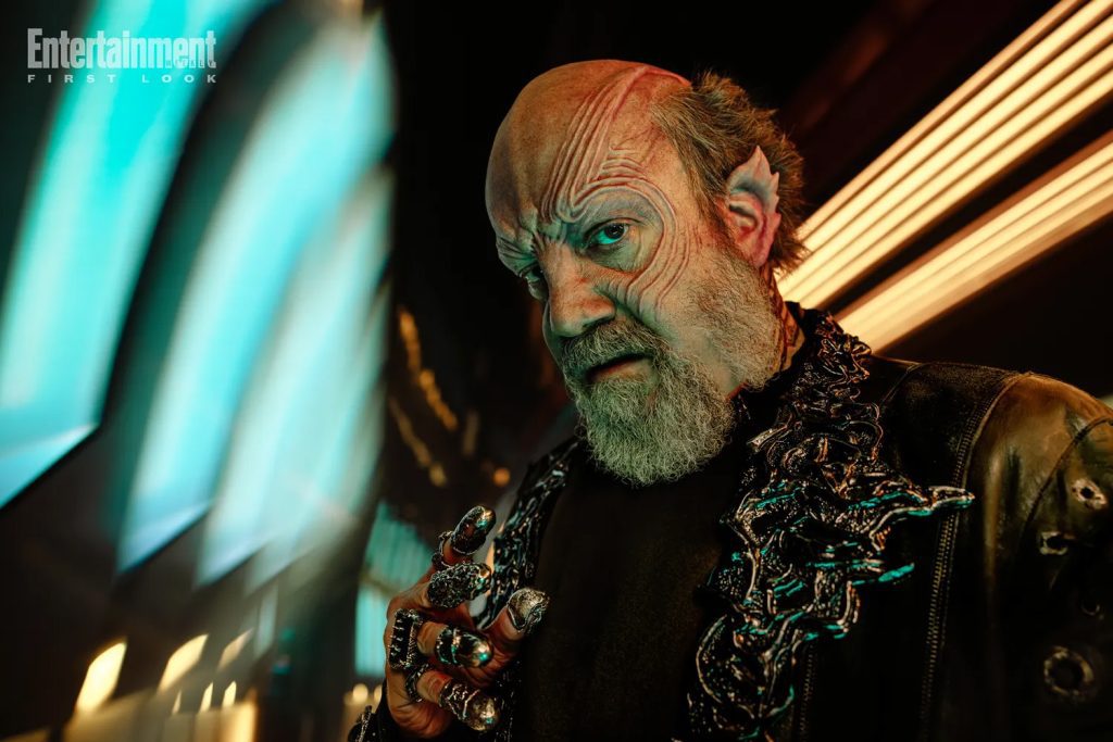 Paul Giamatti con elaborato trucco alieno, indossa una giacca scura e anelli vistosi.
