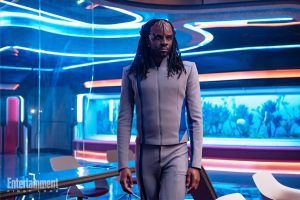 Karim Diane in piedi, con un'uniforme futuristica e capelli lunghi intrecciati, in un ambiente illuminato al neon.