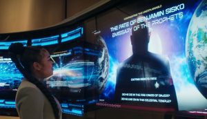 SAM osserva un grande display olografico blu in un ambiente futuristico. Lo schermo mostra testo e immagini relative al destino del personaggio di Star Trek, Benjamin Sisko.
