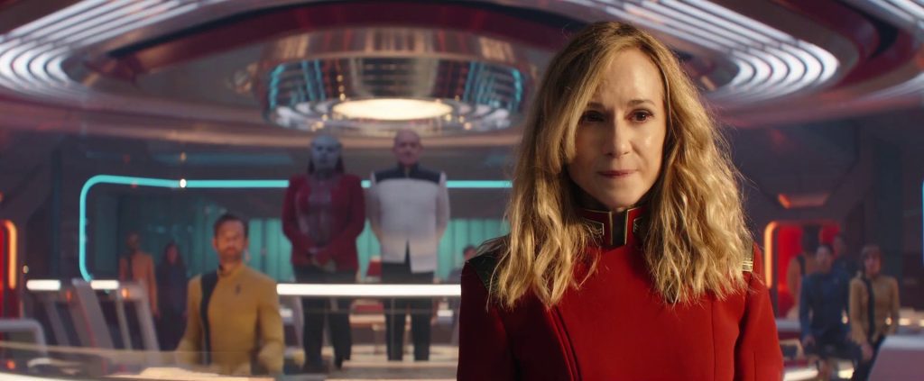 Una donna con capelli biondi di media lunghezza e un'uniforme rossa della Flotta Stellare, con dettagli dorati e verdi sul colletto, guarda con espressione seria e concentrata. Si trova in un ambiente futuristico che sembra il ponte di un'astronave o un'aula dell'Accademia, con luci calde e altri cadetti e ufficiali in uniforme sullo sfondo.